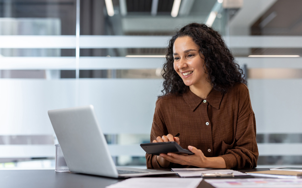 A Smiling Woman Uses A Calculator While Working On A Laptop In An Office Setting