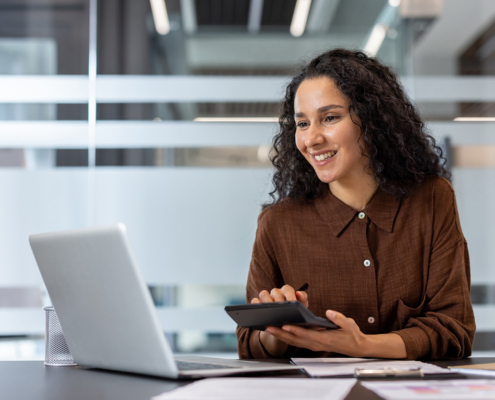 A Smiling Woman Uses A Calculator While Working On A Laptop In An Office Setting